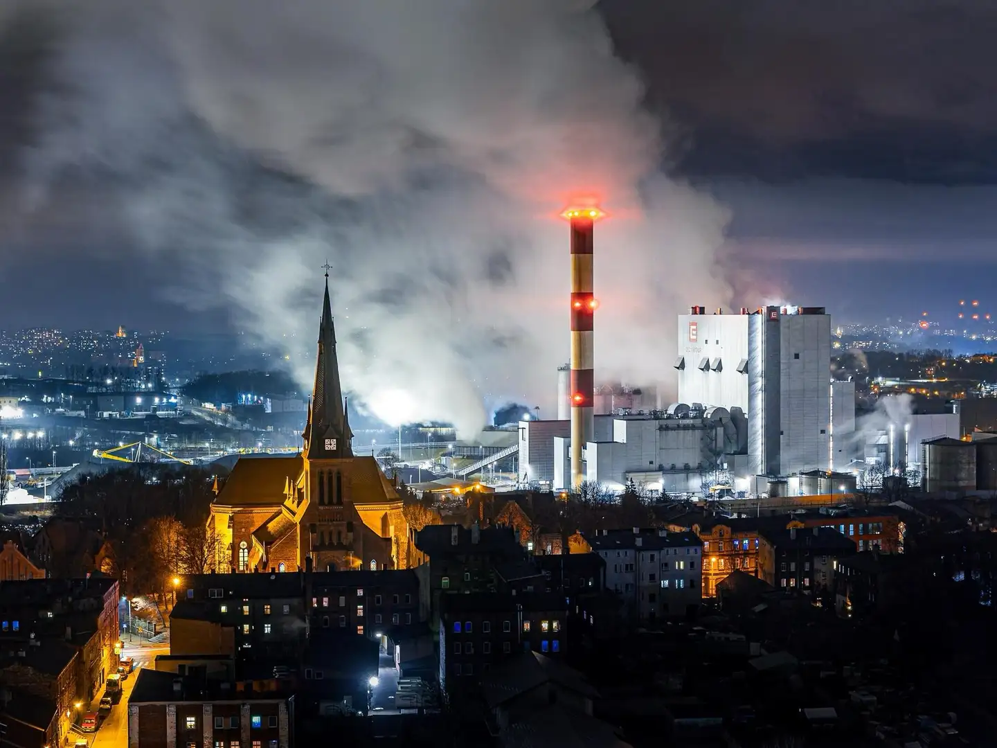 Nocny widok Chorzowa: oświetlony kościół, za nim kominy elektrociepłowni i kłęby dymu